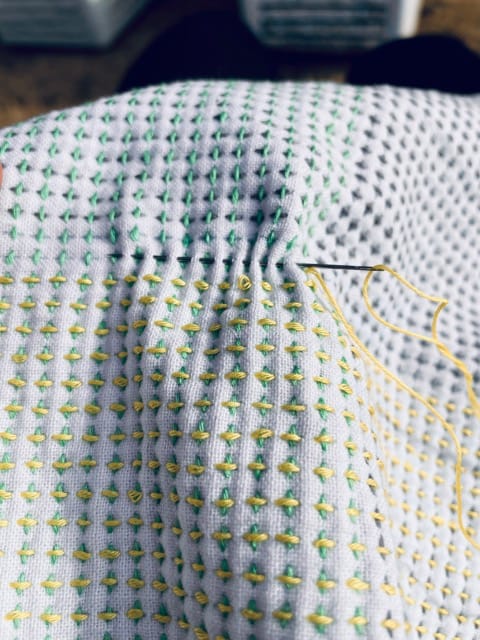 A sashiko kitchen cloth with the jūjizashi pattern in green, yellow and gray. A needle is sticking in the fabric.