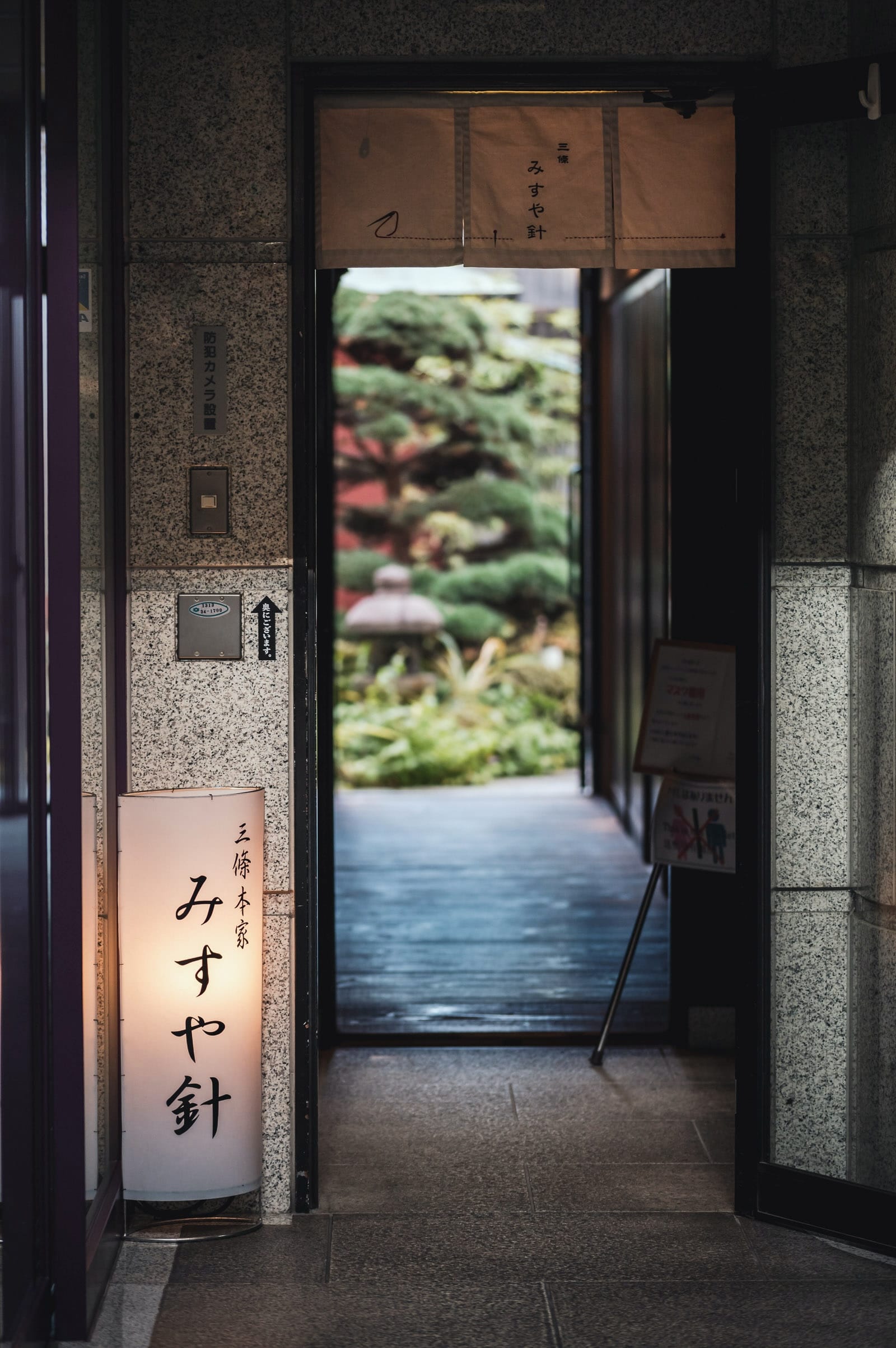 A lit up sign with Japanese writing saying "Misuyabari" in front of an open door leading to a small garden