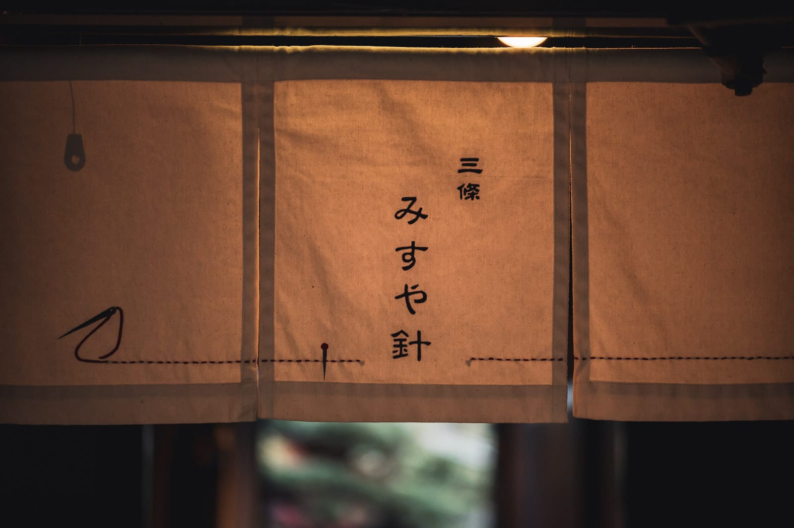 A fabric noren in beige with kanji saying "misuyabari" and a line of thread sewn in marking a needle shop's entrance