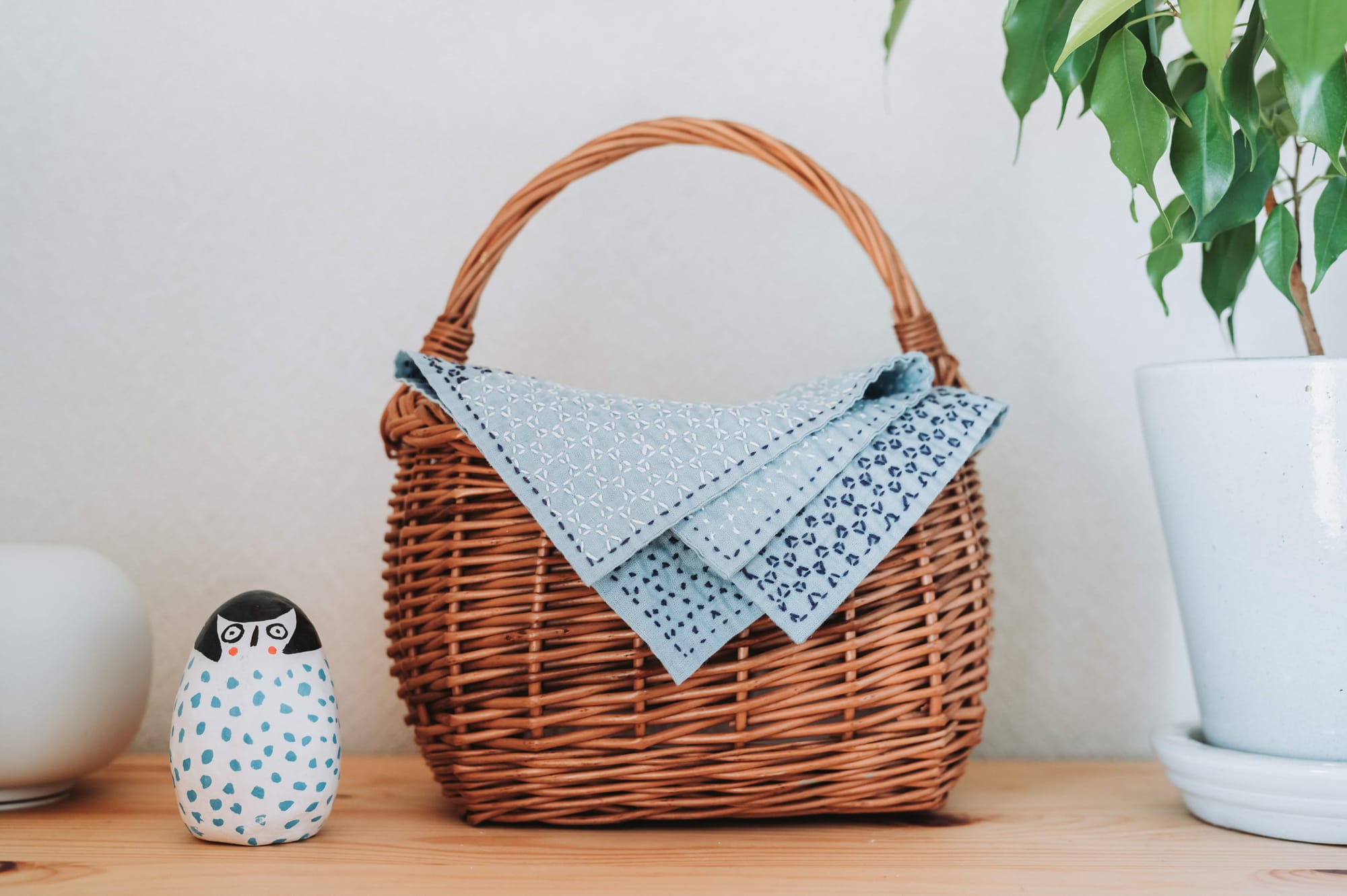 A kagome sashiko pattern kitchen cloth draped across a small basket with a plant and a figurine next to it.